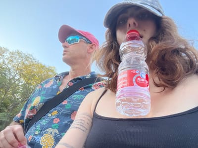 An outdoor close-up shot features two individuals under a clear blue sky. In the foreground on the right, a young adult female, wearing a camouflage-patterned baseball cap and a black tank top with thin straps, holds a small clear plastic water bottle with a red cap and a red-and-white Hebrew label (identifying 'Nevi'ot' mineral water) to her mouth, appearing to drink. Faint, abstract striped tattoos are visible on her left arm. To her left, a middle-aged male wears a red baseball cap, blue reflective sunglasses, and a short-sleeved blue Hawaiian-style shirt adorned with colorful floral and pineapple patterns. A black strap, likely from a bag, crosses his chest. He looks upward and to the left, away from the camera. Green foliage from trees is visible in the background on the left. The scene is illuminated by natural daylight. The Hebrew text on the water bottle confirms the setting is in Israel; however, the precise location within the Brener Regional Council is not discernible from the visual information provided.