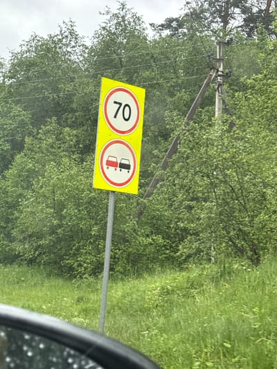The image displays a roadside scene with two traffic signs on a metal pole, set against a backdrop of dense green foliage and trees. The signs are yellow and rectangular, with the upper sign featuring a red circle and the number "70" in black, indicating a speed limit of 70. Below it, the second sign shows a red circle with a diagonal red bar over two car icons, one red and one black, signifying a prohibition of overtaking. A power line pole with insulators is visible to the right of the signs, and the foreground shows the edge of a vehicle, likely a car, with a wet surface reflecting light. The environment is lush and green, suggesting a rural or suburban setting, possibly during or after rain given the wet appearance of the car's surface. The location is identified as Usmangali, Russia.