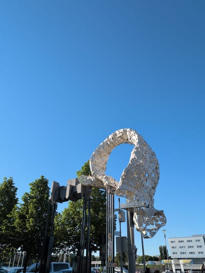 An outdoor, ground-level, medium shot captures a large, abstract metal sculpture against a clear blue sky in Oulu, Finland. The sculpture, composed of numerous irregular, reflective metal pieces assembled into a flowing, organic shape reminiscent of a ribbon or a bird's wing, dominates the foreground. To the left of the main sculpture, a series of dark, angular, geometric metal elements are suspended on vertical poles. Behind these structures are lush green trees. In the background, a white, multi-story building with numerous windows is visible, along with other urban elements like streetlights and signs. Cars and pedestrians are present at the lower edges of the frame, indicating a public space. The overall lighting suggests a sunny day.
