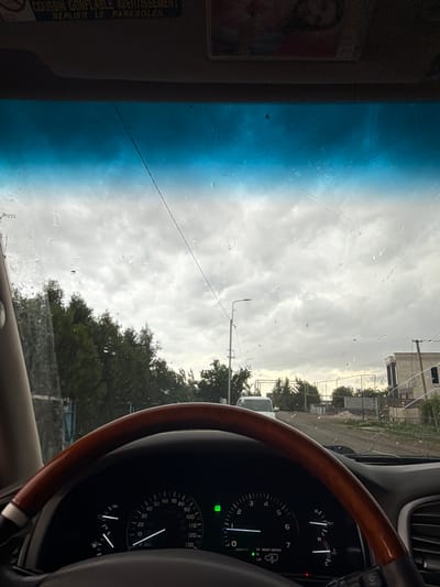 A view from inside a car, likely stationary, with the dashboard, steering wheel, and a portion of the windshield visible. The car appears to be parked on a street in what seems to be a residential or commercial area, with buildings and trees seen through the windshield. Raindrops are visible on the windshield, and there's a crack across it. The dashboard shows a speedometer and other gauges, with a wooden steering wheel in the foreground. A sun visor on the left has a picture and a warning label in French. The exact location in Dimitrov, Armenia cannot be determined from the visual data alone.