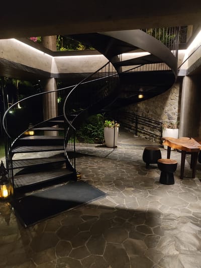 This image depicts a dimly lit outdoor or semi-outdoor common area in Mas, Indonesia, at night. The central feature is a large, black metal spiral staircase with dark wooden treads and a matching metal handrail, ascending towards the upper levels of a multi-story structure. At the base of the staircase, a black rectangular doormat is visible, and a decorative antique-style lantern with a warm glowing light source sits on the flagstone floor beside it.
The floor throughout the scene is paved with irregular, light-colored flagstones. To the right of the staircase, a seating area is arranged, consisting of a rustic wooden table with a natural, live-edge top and three dark, cylindrical stools with wooden seats. Beyond this, a stone-faced wall extends, partially obscuring a darker, possibly sloped pathway or ramp with a low black railing. Two tall, white cylindrical planters containing green foliage are positioned near this wall.
The surrounding environment features concrete columns and walls, some of which are illuminated by horizontal, warm-toned LED strip lights embedded along their edges. Lush green vegetation and darker foliage are visible in the background and upper sections of the scene, indicating an integrated garden or natural setting. No individuals or active subjects are present in the image; the scene is static.