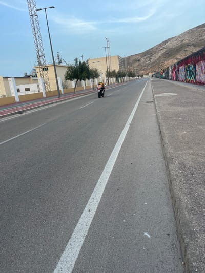 The image depicts a scene along the waterfront in Cartagena, Spain, likely near the port area. A person is riding a motorcycle down a paved road, visible in the center of the frame. The road has a distinct white line marking its center. To the left of the road, there is a raised walkway with a red surface, bordered by a low white and tan wall. Behind this wall, a large industrial building and a tall metal tower with antennae are visible. Trees are planted along the edge of the walkway.
To the right of the road, a concrete sidewalk runs parallel to a graffiti-covered retaining wall. This wall, decorated with vibrant, colorful graffiti, ascends upwards, hinting at the rocky hillside that forms the backdrop of the scene. The sky above is a pale blue with wispy clouds. The overall impression is an urban coastal environment with a mix of industrial structures, public infrastructure, and natural elements.