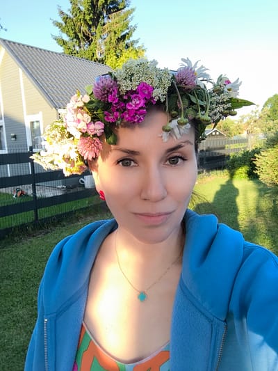 A woman is taking a selfie in a yard with a house and trees in the background. She is wearing a blue hoodie and a flower crown. The woman has dark hair, brown eyes, and is smiling. The house is light-colored with a gray roof. There is a black fence in the foreground. The sky is blue and the sun is shining. The location is Dzērumi, Latvia.