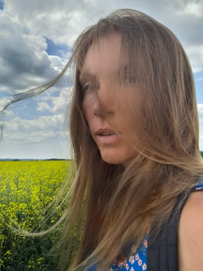 A close-up, medium shot captures a woman with long, blonde hair standing in a field of yellow flowers under a cloudy sky. The woman's face is partially obscured by her hair, and she is looking to the left of the frame. Her mouth is slightly open, and her eyes are not fully visible. She is wearing a blue garment with a floral pattern and a dark backpack. The field of yellow flowers stretches to the horizon, meeting a distant tree line. The sky is a mix of white and grey clouds with patches of blue visible. The image appears to be taken in Treshchevka, Russia, as indicated by the user's prompt.