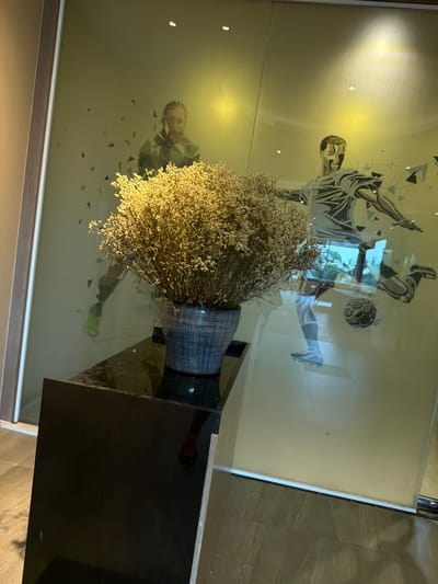 This is a medium shot, eye-level photograph featuring a dark, reflective surface in the foreground, possibly a table or counter, with a ceramic pot containing dried flowers placed on it. The pot is blue and white checkered. Behind the pot, a large glass panel acts as a backdrop, showcasing a graphic design. This design depicts two stylized figures of male soccer players in action, rendered with sharp, angular lines in shades of grey, blue, and white. Geometric shapes, resembling triangles and polygons, are scattered around the players, adding to the dynamic and abstract aesthetic. The lighting is warm and ambient, creating highlights on the glass and the dried flowers. The environment appears to be indoors, possibly a hotel lobby or a modern commercial space in Bang Muang, Thailand, given the architectural style and the type of decorative elements. The overall impression is one of contemporary art and interior design.