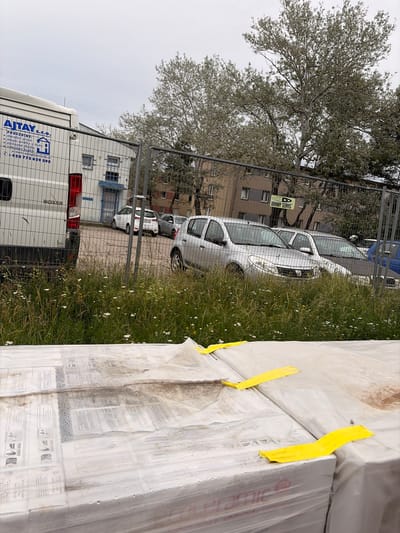 The image shows an outdoor scene in Příbram, Czechia, with a white van on the left, parked in front of a building with a chain-link fence. The van has "AJTAY" and "PROVADIMET" written on its side, along with a phone number. Beyond the fence, several cars are parked, including silver sedans and a dark-colored car. In the background, there are apartment buildings and trees. The foreground is filled with tall grass and wildflowers. In the very bottom of the image, stacked pallets covered in plastic sheeting and yellow straps are visible. The sky is overcast.