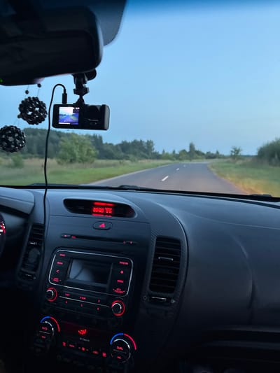 The image shows the interior of a car, looking out at a road in a rural area. The car's dashboard is visible, with the radio and climate controls illuminated in red. A dashcam is mounted on the windshield, and two black, spherical air fresheners hang from the rearview mirror. The road ahead is asphalt with white lines, and it curves to the right. The surrounding environment is green with trees and fields, and the sky is a pale blue, suggesting it is either early morning or late evening. The time displayed on the car's clock is 20:20. The location is identified as Vorotynets, Russia. There are no people visible in the image.