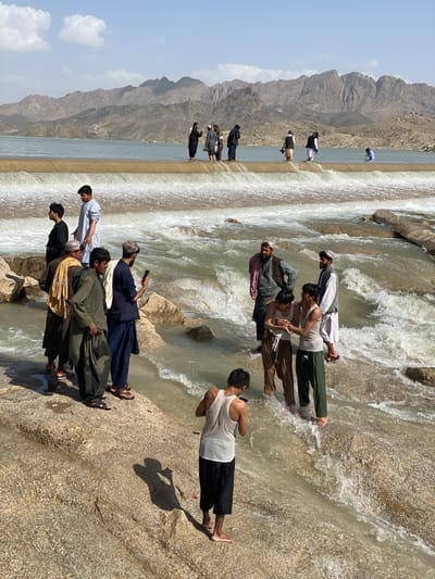 A group of people are gathered at a dam and spillway with a river. Some of the people are standing on the dam, while others are in the river. The people in the river are interacting with each other. The river is flowing rapidly and has whitewater rapids. The background shows a mountain range. The sky is clear with a few scattered clouds. The location is near the city of Kim in Afghanistan.
