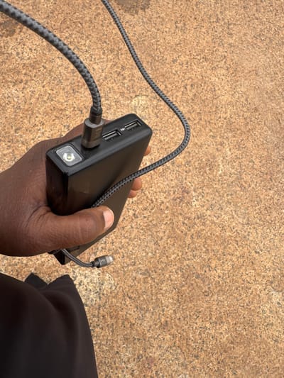 In Jawu, Nigeria, a person's hand holds a black power bank, its charging cable plugged into two USB ports. A bright LED light is visible on the power bank. The background is an outdoor, textured surface, possibly a pavement or ground.