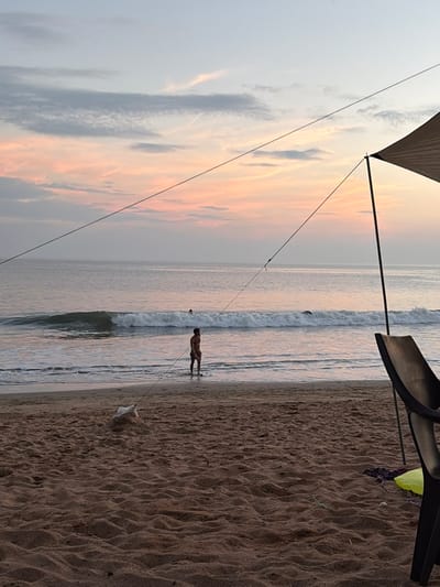 This image displays a coastal scene on a sandy beach in Gokarna, India, observed at either dawn or dusk under a sky featuring pale blue and pink cloud formations. The ocean extends across the midground and background, exhibiting small, breaking waves. An adult male, wearing dark trunks, stands approximately ankle-to-calf deep in the shallow surf, facing towards the right side of the frame. Further offshore, a single individual's head is visible above the water, positioned amidst a breaking wave. The foreground consists of brown, rippled sand. A dark, angular tarp or tent awning with a supporting pole is partially visible in the upper right. Two dark diagonal ropes, identified as guy lines, traverse the frame; one is secured to a sand mound containing a white, possibly weighted, object in the lower center. To the lower right, a dark, curved-back plastic chair is partially visible, alongside a neon yellow object and a dark piece of fabric resting on the sand.