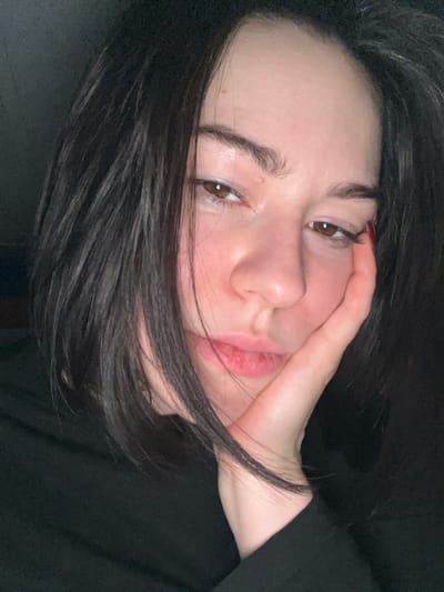 A woman with dark hair and light skin poses with her hand touching her face. She has brown eyes and full lips. Her hair is disheveled, partially obscuring her face. The image is taken at a close-up angle, with the woman's face angled towards the camera. A small glimpse of red nail polish is visible on one of her fingers. The background is dark, creating a high-contrast effect. The overall lighting is dim and moody.