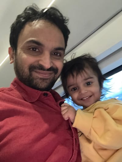 A close-up shot shows a man with dark hair and a beard smiling at the camera. He is wearing a red shirt. A baby, wearing a yellow sweater, is perched on his shoulder. The baby smiles widely with a few visible teeth. The background features a blurred combination of white walls, a door and window with some light streaming through, suggesting an indoor setting. The image is rotated slightly to the left.