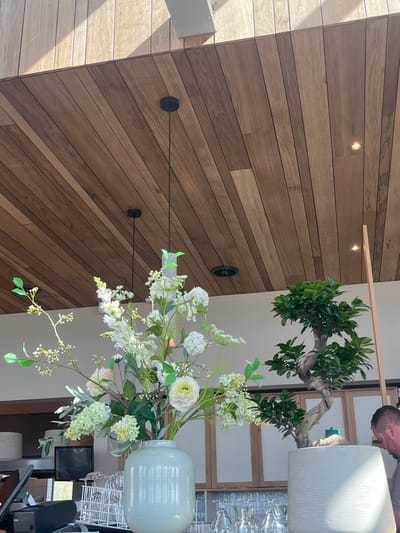 The image presents an interior scene within an establishment located in Overveen, Netherlands. A light greenish-grey cylindrical vase containing a large artificial floral arrangement of white and light green flowers, including ranunculus-like blooms and foliage, occupies the left-center foreground. To its right, a potted bonsai tree with a gnarled, light-colored trunk and green leaves rests in a textured light grey cylindrical pot. Both decorative elements are situated on a light-colored counter. Below the counter, racks holding inverted stemmed glassware are visible. A black point-of-sale terminal is partially visible on the far-left counter. The ceiling is composed of horizontal wooden planks of varying tones, featuring recessed spotlights and two suspended black pendant light fixtures. A dark circular air vent is visible in the upper right section of the ceiling. Natural light enters from an unseen source, casting shadows on the upper left wooden ceiling panels. In the far right background, the right side of a man's head and shoulder are partially visible, showing short dark hair, as he faces away from the camera.