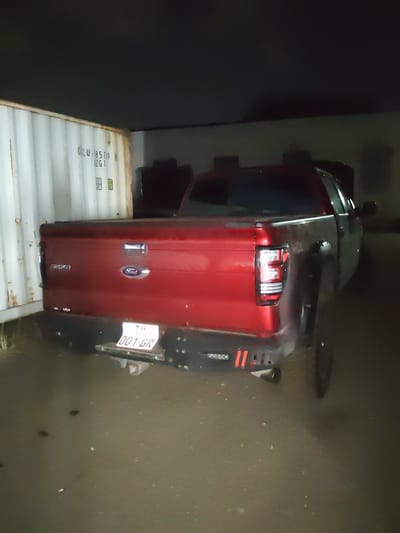 A red Ford F-150 pickup truck is parked in front of a white shipping container at night. The truck's tailgate is visible, displaying "F-150" and the Ford logo. A license plate that reads "001-GR" is attached to the rear bumper. The scene appears to be an outdoor industrial or storage area, with the ground being asphalt or a similar hard surface. The sky is dark, indicating nighttime. The location is identified as Krtsanisi, Georgia.