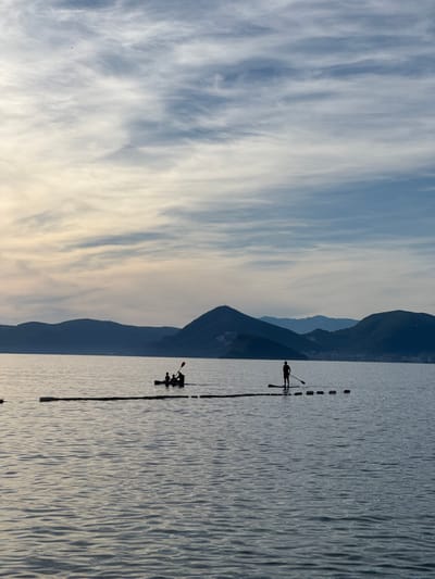 The image depicts a calm body of water with silhouetted figures engaged in water activities against a backdrop of mountains. In the mid-ground, a small boat, possibly a kayak, carries at least three individuals. To the right, a single person stands on a paddleboard, holding a paddle. A line of buoys is visible in the water near the paddleboarder. The water is gently rippled, reflecting the soft light of the sky. The horizon is marked by a range of dark, undulating mountains, some with faint signs of human settlement. The sky is filled with wispy clouds, transitioning from a pale yellow near the horizon to a soft blue higher up. The overall impression is one of tranquility and outdoor recreation in a coastal setting. The image appears to be taken in the vicinity of Rijeka Reževići, Montenegro.