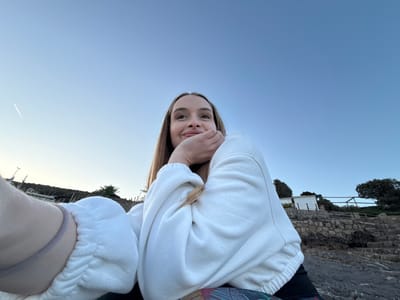 This full-shot selfie captures a young woman looking directly at the camera, holding the device from a slightly lower angle. She has long, straight light brown hair, and her left hand rests beneath her chin, creating a relaxed pose. She wears a white, possibly terry cloth, hooded sweatshirt or jumper, with a dark garment visible near her left shoulder. A grey elastic band is on her right wrist. Her expression conveys a subtle smile and a sense of contentment.
The setting appears to be a coastal or lakeside area in Seoca, Montenegro. Behind the woman, a textured stone wall or rocky terrain is visible, interspersed with dark green foliage and bushes. In the midground, beyond the wall, a distant shoreline stretches across the frame, featuring small white buildings, possibly houses or sheds, and more trees. On the far right, faint structures resembling docks or boats can be seen along the water's edge.
The clear sky dominates the upper portion of the image, transitioning from a deeper blue near the horizon to a lighter, almost white-blue towards the top. A faint, thin white contrail, likely from an airplane, is visible in the upper right quadrant of the sky. The lighting suggests either early morning or late afternoon, with soft, even illumination and no harsh shadows, indicating calm and pleasant weather. There is no visible text in the image.