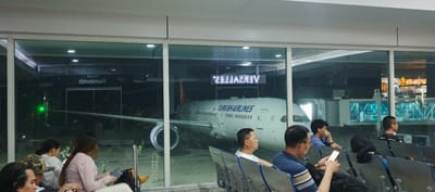 The image shows a waiting area at an airport in Catia La Mar, Venezuela. A Turkish Airlines aircraft is visible through a large window. The word "VERSALLES" is displayed in reverse on a sign above the plane. Inside the waiting area, several people are seated, some looking at their phones or out the window. Other signage says "VIALES Humboldt VIP". The lighting suggests it is nighttime.