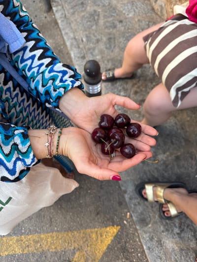 A close-up, high-angle view depicts a scene on a textured gray stone pavement in Deià, Spain. In the foreground, the hands of an individual, wearing a long-sleeved garment with a blue, white, and black zigzag pattern, are cupped together, holding approximately nine dark red cherries with stems. The individual's left wrist displays a stack of bracelets, including a rose-gold chain with star charms, a red string, and a green beaded bracelet, while their fingernails are painted a deep fuchsia. A light-colored bag or garment with a partial green logo is visible below their hands. To the right in the mid-ground, the lower legs of a second individual, wearing a brown and white striped garment and a black slide sandal on their right foot, are present near a black cylindrical bollard. The lower right foreground reveals the foot of a third individual, clad in a gold-colored sandal, with fuchsia-painted toenails. The pavement features a thick, broken yellow painted line in the bottom left and two discarded cigarette butts in the bottom right.