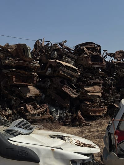 The image displays a vast open-air scrap metal facility located within the Sdot Negev Regional Council, Israel, under a clear, light blue sky. The central feature is an immense, towering mound of severely damaged and corroded vehicle bodies and components, primarily consisting of crumpled car chassis. These vehicles exhibit advanced stages of rust, dismemberment, and crushing, indicating a prolonged period of outdoor exposure and processing for scrap. The pile dominates the midground and background, obscuring the horizon. In the immediate foreground, a dry, dusty, and light brown earth terrain is visible, sparsely covered with small, desiccated plant matter. Partially visible on the left foreground is the front section of a white or light-colored vehicle, featuring a dented hood with significant rust spotting and a black snorkel attachment on its left side. To the right, the rear portion of another vehicle, possibly gray, with a fractured red taillight assembly, is partially observable. No individuals or active operations are present in the scene.