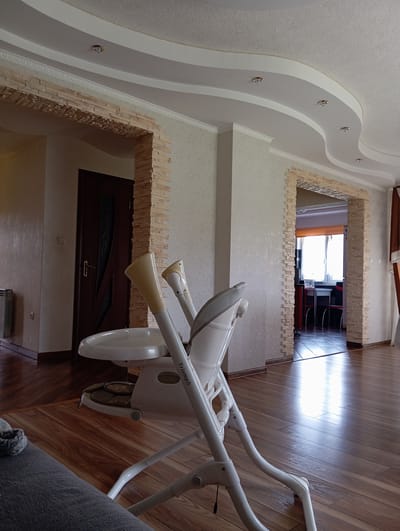 In the city of Hlynytsi, Ukraine, an indoor scene captures a modern living space. The room features a curved ceiling with recessed lighting, complemented by decorative plasterwork and stone veneer accents around archways. The flooring is polished hardwood. In the foreground, a white high chair with a beige seat is positioned, facing towards the viewer. A dark wooden door is visible in the left background, and a doorway to another room is in the right background, revealing a glimpse of a dining area with a table and chairs. The scene appears to be a domestic environment, possibly a dining or living area.