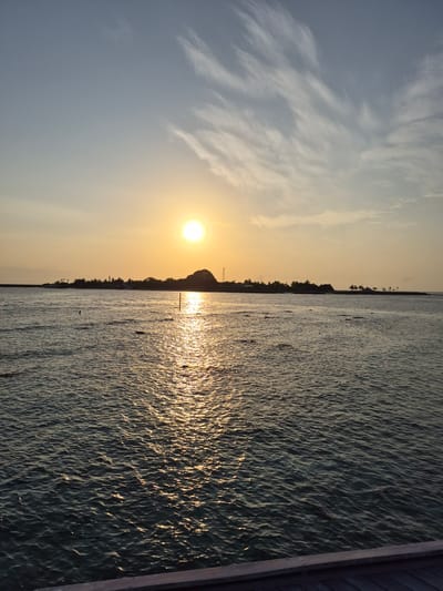 The image depicts a sunset over the ocean near Bodufolhudhoo, Maldives. The sun is low in the sky, casting a golden glow on the water's surface and reflecting off the waves. The sky is a mix of light blue and yellow, with wispy clouds. The shoreline is visible in the distance, with vegetation. A wooden structure is at the edge of the water on the right.