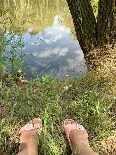The image depicts a low-angle, point-of-view shot of a person's feet wearing pink slides, resting on grassy terrain next to a body of water. The water, which appears murky and reflects the sky and surrounding trees, suggests a natural setting, possibly a riverbank or pond edge. A large tree trunk, partially covered in moss or lichen, stands to the right of the frame, its rough bark textured by sunlight. The foreground is dominated by lush green grass and dried vegetation, indicating a rural or natural area. The scene is bathed in natural light, creating shadows and highlighting the textures of the grass and tree bark. There is no indication of urban infrastructure or any specific landmarks that would definitively place the scene within the city of Ust'-Dzhigutinka, Russia.