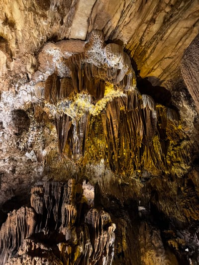 This is a photograph taken inside Tham Phu Kham, a cave located near the city of Vang Vieng in the Vientiane Province of Laos, not Nadao. The image depicts the interior of the cave, showcasing a variety of speleothems, which are natural formations created by mineral deposits.

The ceiling of the cave is heavily adorned with stalactites, which hang downwards like icicles. These formations vary in size, shape, and color, ranging from rough, textured grey and brown structures to smoother, elongated formations with a yellowish-orange hue, likely due to mineral content and lighting. In some areas, particularly towards the center and right of the image, the stalactites are densely packed and illuminated by artificial lights, highlighting their intricate details and creating dramatic shadows.

The walls and ceiling of the cave exhibit a rugged, uneven texture, with layered rock formations and evidence of mineral growth. There are also areas of lighter colored mineral deposits, possibly calcite, that appear as white or pale grey crusts on the rock surfaces.

The overall environment is dark and cavernous, with the illumination focusing on the speleothems, creating a surreal and geological display. No people or animals are visible in the photograph. The image is a close-up view, emphasizing the textures and forms of the cave's natural formations.