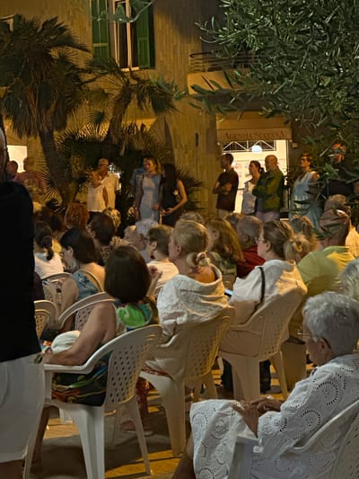 In Ospedaletti, Italy, an outdoor evening gathering is underway, with a crowd of approximately 30-40 individuals seated on white plastic chairs arranged in rows. The event appears to be a performance or presentation, as many attendees are facing forward, towards an unseen focal point. The background features a building with a sign reading "AGENZIA SULTANA," indicating a commercial establishment, possibly a real estate agency. Palm trees and olive branches frame the scene, suggesting a Mediterranean setting. The lighting is warm and artificial, characteristic of evening events. The attire of the attendees varies, from casual to slightly more formal, with some individuals wearing light-colored clothing. The mood seems relaxed and engaged, with people observing, listening, and interacting amongst themselves.