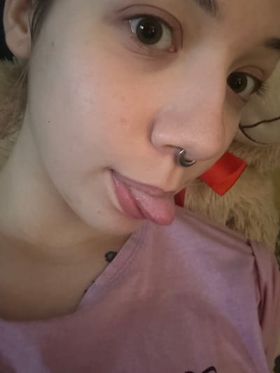 A close-up selfie of a young person with dark brown eyes and light skin is shown from the chest up. The person has a septum piercing and is sticking out their tongue, which has a pink gloss on it. They are wearing a pink t-shirt and have a small, dark tattoo on their collarbone. In the background, a fluffy teddy bear with a red ribbon around its neck is visible. The image does not contain information about the location in the city of Olaine, Latvia.
