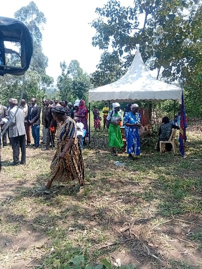 An outdoor daytime gathering is depicted in Butali Chegulo ward, Kenya. Approximately 25 individuals, primarily adults, are distributed across an unpaved, sparsely vegetated area. In the foreground, a woman wearing a black, brown, and gold patterned dress with light green shoes walks towards the left, her left arm slightly bent. To her left, a cluster of approximately ten men, several in suits or collared shirts, stand facing the center-right. A dark, metallic object, consistent with video camera equipment, is partially visible in the extreme left foreground. A white, pointed-roof pop-up tent occupies the mid-ground on the right. Beneath the tent, a dark table holds unidentified objects, and a person is seated on a light-colored plastic chair. Several women, attired in colorful dresses and head coverings (including green, blue, and patterned fabrics), are positioned near and under the tent. A multi-colored flag, predominantly red and blue, is mounted on a pole immediately to the right of the tent. The background features a clear sky above diverse tree lines, with tall, slender trees on the left and denser, broader-leafed trees on the right.