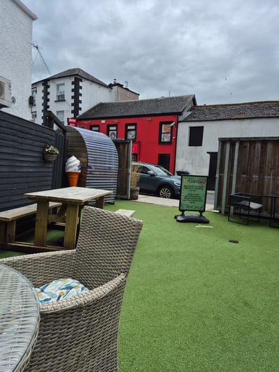 This image depicts an outdoor seating area, likely in Carrick-On-Shannon, Ireland, featuring artificial turf flooring. In the foreground, a wicker chair with a patterned cushion sits beside a glass-topped table. A wooden picnic table with benches is positioned further back. To the left, a dark wooden fence encloses the area, with a hanging basket containing greenery attached to it. An oversized ice cream cone decoration stands near a curved corrugated metal structure.
In the background, several buildings are visible. One building is painted bright red with black trim and features windows and signage, including "APACHE OPEN". Adjacent to it is a white building with black architectural details. A dark-colored car is parked on the street in front of these buildings. A green A-frame sign on the artificial turf reads "Kierans Gala Welcome to Gala Garden Seating area. Only food purchased on these premises can be consumed here. Thank You". Wooden fencing separates different sections of the outdoor space, and some potted plants are present. The sky is overcast with clouds.