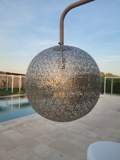 The image, viewed upright, features a large, spherical, metallic lamp shade or decorative orb with an intricate, organic, laser-cut pattern, centrally positioned and suspended from a dark, curved metal arm. The metallic surface reflects warm, golden light, suggesting an internal light source or the sun's low angle.

The background reveals a clear, light blue sky with minimal cloud cover in the upper left. Below and to the right of the sphere, a light-colored paved surface, likely a patio or poolside, is visible. Adjacent to the paving is a tranquil swimming pool reflecting the sky and distant architectural elements, possibly white pergolas or structures, along with green foliage. The reflections in the water also show the warm, golden light. A partial view of a white, modern outdoor furniture piece is visible on the far right.

The overall lighting indicates late afternoon or early evening, characterized by the soft, warm glow. The scene is static, depicting no human activity. The setting appears to be a peaceful outdoor area, possibly a resort or a private residence in Sogliano Cavour, Italy, featuring decorative lighting and a pool. There is no visible text in the image.
