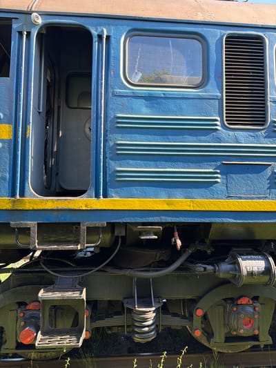 This is a close-up, vertically oriented photograph of the exterior of a blue train car, focusing on its mechanical components and a section of the blue and yellow painted body. The scene appears to be outdoors, possibly in a rail yard or industrial area, given the presence of overgrown vegetation on the right side of the frame. The lighting suggests it is daytime, with strong sunlight casting distinct shadows, indicating clear weather.
The left side of the image displays a portion of the train car's blue bodywork. Prominent features include a rectangular ventilation grille with thin, dark slats positioned in the upper section, and a small, rectangular window with a somewhat reflective glass surface below it. The blue paint shows signs of wear, with some chipped areas visible, particularly around the window and the large, rectangular opening below it. This opening reveals an interior space of the train, also painted blue, with a smaller, dark rectangular panel visible within. Vertical ridging is a dominant textured element on the blue bodywork, running from near the top of the frame down to the large opening. A thick, bright yellow stripe runs vertically along the edge where the train car's body meets its mechanical undercarriage.
The right side of the image is dominated by dark-colored, heavy mechanical parts of the train's undercarriage, likely part of the suspension or braking system. This includes large, circular or semi-circular components, some with bright red accents, possibly indicating critical points or safety features. A prominent cylindrical object, part of what appears to be a pneumatic or hydraulic system, is visible in the upper right quadrant. This cylinder has white text on it, reading "ПРОГРЕСС" and "271(10)25". There are various hoses, cables, and structural supports connecting these mechanical elements. The overall impression is one of robust, industrial machinery.
There are no people visible in the frame. The focus is solely on the static elements of the train and its machinery. The location context is Nizhnyaya Maktama, Russia.
