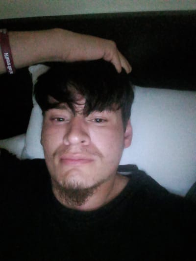 A medium-shot photograph shows a person with short, dark hair, brown eyes, and a mustache and goatee. The person is wearing a black shirt and is lying on a white pillow. A hand rests on the person's head, and a red wristband is visible on the wrist. The location cannot be determined from the image.