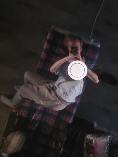 The image captures an indoor scene from a high-angle perspective, showing an individual lying on what appears to be a bed or mattress. The person is holding a smartphone, seemingly taking a selfie or recording themselves, with their face obscured by a prominent white circular object. They are wearing a light-colored top, possibly light blue or grey, and light brown or beige trousers. Their hair appears to be light brown or blonde.
The surface beneath them is covered with a dark plaid fabric, patterned with purple and black checks. In the upper right corner, a dark pillow with a distinct red floral design is partially visible. The surrounding area, likely a floor or another dark surface, is dimly lit, suggesting it might be evening or a poorly lit room. A thin, dark cable or wire runs across the dark background on the left side. The setting is characteristic of a personal living space or bedroom in Nadym, Russia. No visible text is present in the image.
