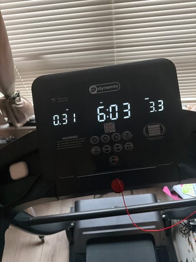 Here is a forensic description of the image:
The image shows a close-up of the console of a Dynamix treadmill. The console is black and features a digital display showing a speed of 3.3, a time of 6:03, and a distance of 1.81. Several buttons are visible, including speed controls, program selection, and mode selection. A safety key attached to a red cord is also visible. The background includes a window with closed vertical blinds and a person with fair skin tying the lace of a shoe. The environment appears to be indoors. There is no information to identify this image's location in the city of Enniskillen, United Kingdom.