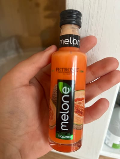 A hand is holding a small, clear glass bottle filled with a pale orange liquid. The bottle has a black cap and a label with a black band. On the label, in white text, is the word "melone". Above this, in smaller text, is "PETRONIO" and "CREMA LIQUORE". To the right of "melone", on a green background, is the word "liquore". The bottle is lying horizontally, held by the fingers of a person's hand, with the thumb supporting the underside. The background is blurred and appears to be a white, textured surface, possibly a shelf or table, with a suggestion of other objects in the distance. The lighting is bright, indicating daytime, and the focus is on the bottle and the hand holding it. No specific weather or emotional cues are evident.