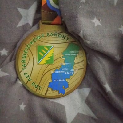 This media file is a close-up, overhead shot of a gold-colored medal lying on a grey fabric background with white stars. The medal is round and features a relief design. The outer edge of the medal has text inscribed in green lettering that appears to read "100 ЛЕТ ТАЙШЕТСКОМУ РАЙОНУ," which translates to "100 Years of Tayshet District." The central part of the medal depicts a map of the Tayshet District. A coat of arms, featuring green and yellow elements, is placed on the left side of the map. Various locations within the district are marked on the map with dots and corresponding Russian names, including "ПОЛИНЧЕТ," "НОВОБИРЮСИНЕЯ," "ЩИТКИНО," "ЮРТЫ," "КВИТОК," "БИРЮСИНСК," "ТАЙШЕТ," and "СОЛЯНАЯ." The medal appears to be a commemorative item, likely related to an anniversary celebration for the Tayshet District in Russia. The fabric background suggests the medal is resting on a blanket or textile. There are no people visible in the image, and the focus is solely on the medal and its details.
