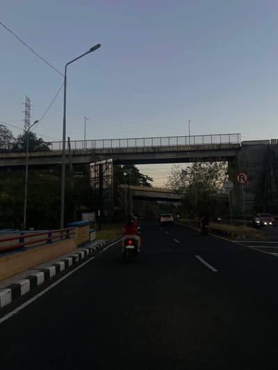 The image depicts a twilight scene on a road in Maumbi, Indonesia, near a bridge structure. A motorcycle carrying two individuals is traveling away from the viewer on the right side of the road. The rider, wearing a red shirt, is visible, and the passenger is partially obscured. The road has visible lane markings, and there are other vehicles on the road, including a white SUV and a red car. A "no U-turn" sign is visible on the right side of the road. The environment includes trees and vegetation on the left and right sides of the road, and a large concrete overpass runs across the upper portion of the image. A utility pole with wires is also present. The sky is clear and shows the transition from day to night.