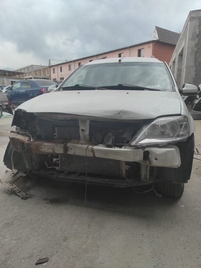 A white car, likely a Dacia Logan, is parked outdoors. It appears to have sustained significant front-end damage, with the bumper removed and the hood dented. The radiator and other engine components are exposed. The car is on a paved surface that resembles a street or parking lot. In the background, there are buildings under construction, with exposed brickwork and scaffolding. Other cars are parked nearby, including a blue sedan. The sky is overcast with clouds. The overall impression is of a car in a state of disrepair or undergoing repairs in an urban or semi-urban environment. The image does not contain any information that specifically places it in Qairat, Kazakhstan.