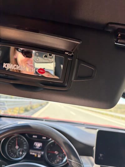 The image shows the interior of a car, specifically the driver's side. The sun visor is pulled down, revealing a mirror with the Cyrillic inscription "КРАСИВА СИ" and a red lipstick mark. The reflection in the mirror shows a person holding a smartphone. The dashboard, steering wheel, and partially visible instrument panel are visible. The speedometer reads 148. Outside the car, a road and vegetation can be seen. There is no definitive information to confirm the specific location in Mikrevo, Bulgaria, but based on the road scene, it could be a local road outside the city of Mikrevo, Bulgaria.