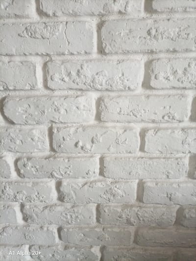 This is a close-up, eye-level, vertical shot of a white painted brick wall. The bricks are laid in a standard running bond pattern, with mortar lines visible between them. The surface of the bricks is rough and textured, with some imperfections and variations in the paint application, suggesting they may be real bricks or a realistic imitation. The lighting appears to be uniform, casting no distinct shadows and highlighting the texture of the bricks. There are no people, actions, or other objects visible in the frame. The image is consistent with a detail shot of interior or exterior wall cladding. Without further context, the specific location in Улукаўскі сельскі Савет, Belarus cannot be determined.