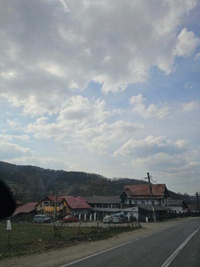 The image depicts a rural or semi-rural roadside scene in Bălteni, Romania, under a partly cloudy sky. The upper two-thirds of the frame are dominated by a sky with numerous white and grey cumulus clouds interspersed with patches of light blue sky.
In the foreground, a two-lane asphalt road extends from the lower right towards the center, featuring a solid white line on its right edge and a broken white centerline further left. To the immediate left of the road is a dirt and grassy verge with sparse, dry vegetation. A small, irregularly shaped white object is visible on the far left of this verge.
The midground features a cluster of residential and possibly commercial buildings, primarily characterized by red or reddish-brown tiled roofs. From left to right: a structure with a red roof and a prominent yellow upper facade is visible. Adjacent to it, a light-colored minivan or van is parked. Next, a red sedan is parked, followed by a row of three light-colored cars, possibly sedans or compact SUVs, parked parallel to the road in a paved area. Further right, a larger multi-story building with a dark lower section, white upper walls, dark wooden trim, and a red tile roof stands prominently. Several utility poles with overhead power lines are visible along the road, particularly on the right side.
The background consists of forested hills or mountains, appearing dormant or in early spring, extending across the horizon behind the structures. No individuals are visible, and no specific actions or interactions are depicted within the scene.