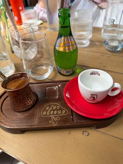 The image depicts a wooden tray on a table. On the tray, there is a small copper pot filled with coffee, and next to it, an empty white espresso cup with a red saucer. The cup has the text "caffè alberto" printed on it, along with a small logo of a bear. In the background, there are several glasses, a green glass bottle with a blue and yellow label, and a small candle in a glass holder. A person wearing a pink and white plaid shirt is partially visible in the background. The overall setting appears to be a cafe or restaurant.
While the image contains elements commonly found in cafes, there is no specific visual information that allows for the determination of its location within the city of Drama, Greece.