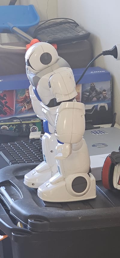 The image depicts a white and blue robot toy, possibly a humanoid or semi-humanoid model, standing on a dark plastic surface. The robot is positioned in a profile view, facing to the right. Its body is segmented into various parts, including a head with a circular black eye, shoulders, arms, a torso, and legs with feet. The robot has a small red accessory attached to the top of its head.
In the background, there are several objects that suggest a cluttered indoor environment. To the left, a blue plastic shovel with a metal handle is visible. Behind the robot, there's a black keyboard, and further back, a blue box with "SONY" printed on it, which appears to be a PlayStation 5 game console box, featuring imagery of characters from a video game. To the right of the robot, another electronic device, possibly a printer or a gaming console, is partially visible. The overall setting appears to be a room with various electronic devices and household items. The lighting suggests an indoor scene, potentially during the daytime. The image does not provide any direct information about the location being Paralimni, Cyprus.