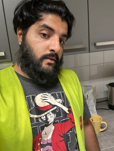 A medium shot captures a man with dark hair, a full beard, and brown eyes wearing a grey t-shirt with an anime character print and a bright yellow vest. He is standing in a kitchen, with grey cabinets and a tiled backsplash visible in the background. A white paper bag and a yellow mug are on the counter. The man is looking towards the camera with a neutral expression. The scene appears to be indoors, likely a domestic setting in Schweitenkirchen, Germany.
