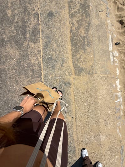 An overhead shot shows a person's legs and torso as they walk along a sandy path adjacent to a concrete walkway. The person is wearing dark brown shorts, a smart watch on their left wrist, and white athletic shoes. They are carrying a brown paper shopping bag with blue lettering that reads "PRIMARK." The environment appears to be a public walkway, possibly near a beach, with sand encroaching on the concrete. The scene is lit by bright sunlight, casting a shadow to the left of the person's feet. The walkway is made of concrete slabs with visible aggregate and is partially covered in sand and debris. The location is identified as Bournemouth, United Kingdom.