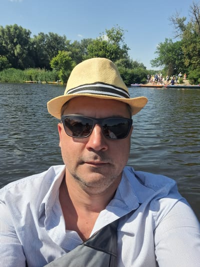 The image shows a man wearing a straw hat and sunglasses, taking a selfie on a body of water. In the background, there is a shoreline with trees and a group of people on what appears to be a beach, some of whom are on paddleboards or kayaks. The water is calm with ripples, and the sky is clear and blue. The man is wearing a light-colored, striped collared shirt. The location is identified as Potoky, Ukraine.