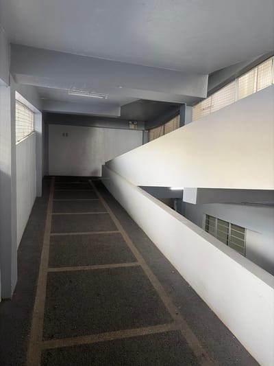 The image depicts the interior of a concrete ramp, likely within a parking garage or a multi-level building, in Dagupan City, Philippines. The ramp is paved with dark asphalt marked with lighter colored lines, creating a grid pattern. The walls and ceiling are painted in various shades of gray. A white concrete barrier runs along the right side of the ramp, separating it from a lower level that features a window with multiple panes. On the left side, a gray concrete wall is visible, with a metal fence or railing running along its top. The environment is brightly lit by artificial lighting, with a fluorescent light fixture visible on the ceiling in the background. There are no people or animals present in the image. The overall scene is functional and utilitarian, characteristic of an urban infrastructure.