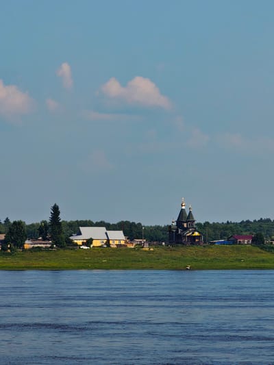 The image depicts a panoramic view of a village located on the banks of a wide river, likely in the city of Potapovo, Russia. The scene is dominated by a bright blue sky with scattered, fluffy white clouds. In the foreground, the choppy surface of a deep blue river fills the lower portion of the frame, reflecting the sky and suggesting a slight breeze.
The middle ground showcases a grassy embankment rising from the river. Several structures are visible on this elevated ground. To the left, a long, low yellow building with a light-colored roof is situated. A tall, dark green evergreen tree stands beside it, and a smaller evergreen is visible further left. A white car is parked in front of the yellow building.
Further to the right, a prominent dark wooden church with multiple golden domes stands out. The church features a complex architectural design with a steep roofline and intricate detailing. Adjacent to the church, a small building with a red roof and a bright blue structure are also present. A dense line of green trees forms a backdrop behind the village, extending across the horizon. The overall impression is of a serene, rural landscape on a clear day.